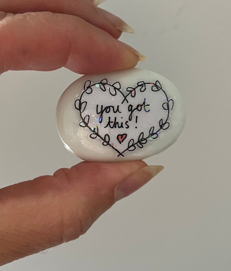 May include: White heart-shaped stone with black ink lettering that reads "You got this!" surrounded by a black floral design. A small red heart is drawn at the bottom of the stone.