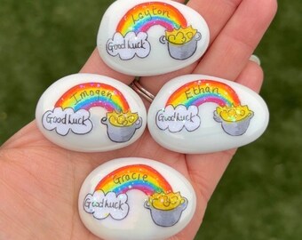 May include: Four white painted rocks with rainbows, pots of gold, and the names Layton, Imogen, Ethan, and Gracie. Each rock also has the words "Good luck" written on it.