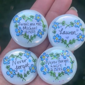 May include: Four round, white glass stones with hand-painted designs. Each stone features blue forget-me-not flowers and green leaves. Text includes "Forget me not Michael 12/5/25", "Lauren", "Never forget", and "Never forget Lucie 3/5/25".