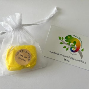 Może przedstawiać: Mały biały woreczek z organzy wypełniony ż&oacute;łtym papierem jedwabnym. Papier jedwabny ma brązową okrągłą naklejkę z tekstem "Handmade with love" czarnym drukiem. Biała karta z ilustracją zielonego kameleona i tekstem "Calm Chameleon Creations Handmade Positive Pebbles and Story Stones" znajduje się w tle.