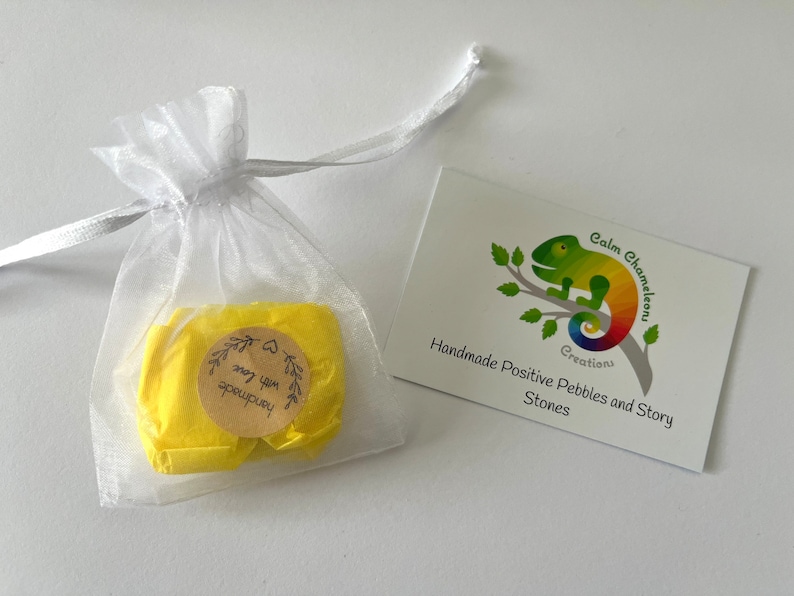 Może przedstawiać: Mały biały woreczek z organzy wypełniony ż&oacute;łtym papierem jedwabnym. Papier jedwabny ma brązową okrągłą naklejkę z tekstem "Handmade with love" czarnym drukiem. Biała karta z ilustracją zielonego kameleona i tekstem "Calm Chameleon Creations Handmade Positive Pebbles and Story Stones" znajduje się w tle.