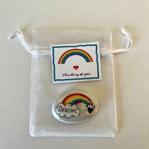 Może przedstawiać: Biały, błyszczący, pomalowany kamień z tęczą i chmurą. Chmura ma napisane na niej słowo "Deacon". Kamień znajduje się w białym woreczku z organzy z kartką, na kt&oacute;rej napisano "Thinking of you" i znajduje się tęcza z czerwonym sercem w środku.
