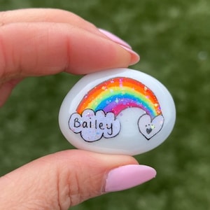 Może przedstawiać: Biały pomalowany kamień z tęczą, chmurą z napisem Bailey i sercem z odciskami łap.