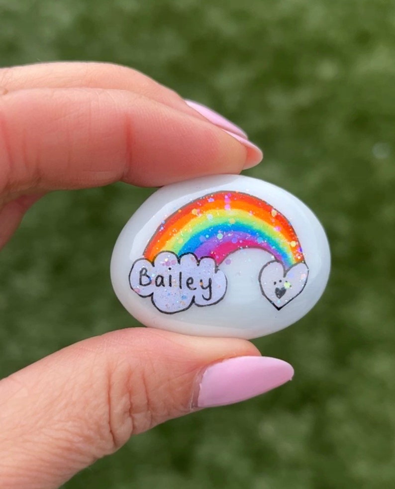 Może przedstawiać: Biały pomalowany kamień z tęczą, chmurą z napisem Bailey i sercem z odciskami łap.