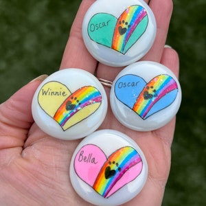 May include: Four painted heart-shaped stones with rainbow designs and paw prints. The stones are white with a glossy finish. The names "Oscar", "Winnie", "Oscar", and "Bella" are written on the stones.