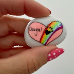 May include: A white heart-shaped stone painted with a rainbow and paw prints. The name "Dougal" is written on the stone.
