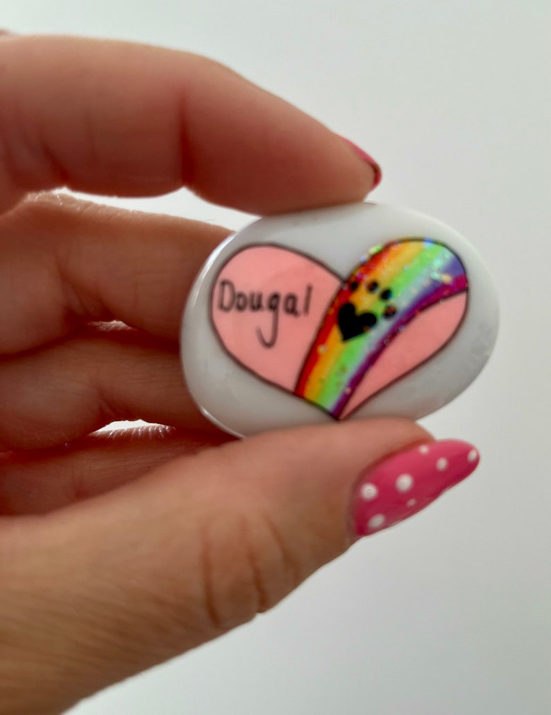 May include: A white heart-shaped stone painted with a rainbow and paw prints. The name "Dougal" is written on the stone.