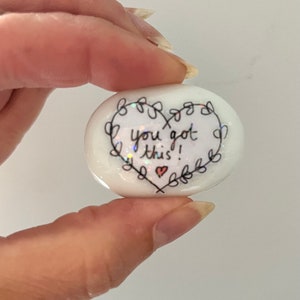 May include: White heart-shaped stone with a black outline and the words "You got this!" written inside. A small red heart is drawn below the text.