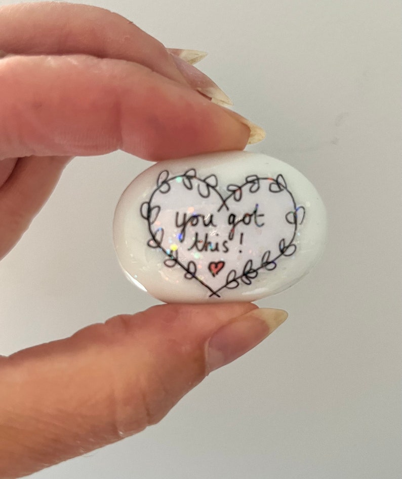 May include: White heart-shaped stone with a black outline and the words "You got this!" written inside. A small red heart is drawn below the text.