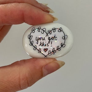 May include: White heart-shaped stone with glitter and black lettering that reads "you got this!" with a small heart drawn next to the text.