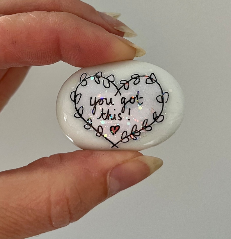 May include: White heart-shaped stone with glitter and black lettering that reads "you got this!" with a small heart drawn next to the text.