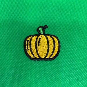 May include: Embroidered patch of a yellow pumpkin with a black outline and green stem, on a green background. The patch is likely for clothing or accessories. The design is simple and suitable for autumn or Halloween.
