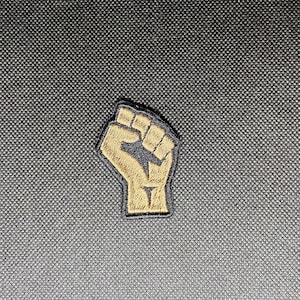 Pode incluir: Um punho levantado marrom com contorno preto, símbolo de solidariedade e resistência. O punho é bordado em um tecido cinza escuro.