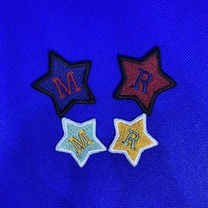 以下が含まれることがあります： 異なる色の「M」と「R」の文字が刺繍されたフェルトの星が4つ。星は青い背景の上にあります。