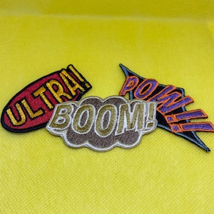 May include: Embroidered patches with comic book style text. The patches include the words "ULTRA!", "BOOM!", and "POW!" in bold, colorful lettering. The patches are on a yellow background.