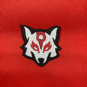 Puede incluir: Parche bordado de cabeza de lobo blanco con detalles rojos y negros. El lobo tiene un diseño estilizado con un círculo en el centro de su frente.