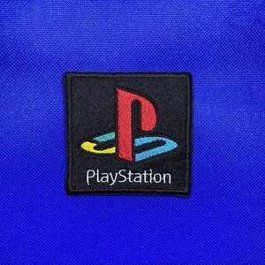 Puede incluir: Un parche negro y blanco con el logotipo de PlayStation, con un diseño rojo, amarillo y azul. El parche tiene la palabra "PlayStation" escrita en blanco debajo del logotipo.