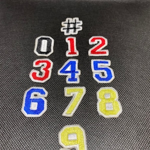 Puede incluir: Conjunto de diez parches termoadhesivos con los números del 0 al 9 y un signo de número. Los parches son blancos con bordados de colores. Los números 0, 1 y 2 son rojos, 3 y 4 son azules, 5, 6 y 7 son amarillos, 8 y 9 son verdes y el signo de número es negro.