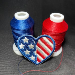 May include: Two spools of thread, one red and one blue, with a heart-shaped patch featuring the American flag design.