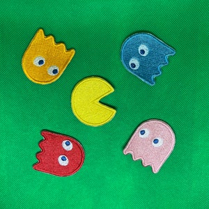 May include: Five embroidered iron-on patches featuring Pac-Man characters. The patches are yellow, blue, pink, red, and orange. Each patch has a different design, including Pac-Man, ghosts, and a Pac-Man with a mouth open.