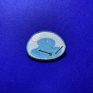 May include: A blue embroidered patch of a cartoon character inside a bubble. The character is blue and has a white outline. The character is holding a pair of chopsticks.