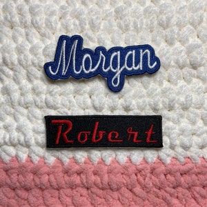 May include: Two embroidered patches with names. The top patch is blue with white stitching and says "Morgan". The bottom patch is black with red stitching and says "Robert".