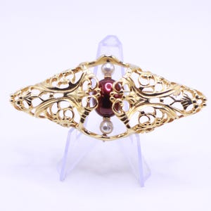 Può includere: Spilla dorata con un design traforato ornato. Presenta una perla centrale rosso scuro e due perle più piccole simili a perle. La lavorazione del metallo a filigrana crea una forma simmetrica ed elegante, adatta alle occasioni formali. Misura circa 8 cm.