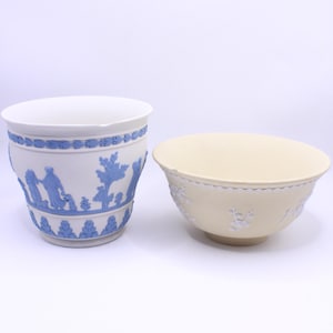May include: Two ceramic decorative pieces. A white planter with blue relief figures and a blue border. Next to it is a cream-colored bowl with a floral relief design around the rim. Both pieces are on a white background.
