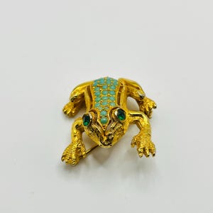 Puede incluir: Broche de rana dorada con detalles turquesa y verde. La espalda de la rana está adornada con pequeñas piedras turquesas, y sus ojos son verdes. El broche tiene un cierre de alfiler.