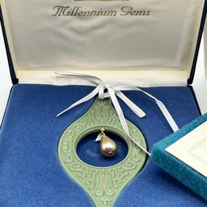 Könnte beinhalten: Ein Wedgwood Millennium Gems Ornament in einer blauen Box. Das Ornament ist hellgrün, tropfenförmig und hat einen goldfarbenen Anhänger. Die Box hat das Wedgwood-Logo und die Aufschrift "Millennium Gems". Ein weißes Band ist befestigt.