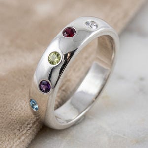 May include: A silver ring with a polished finish, set with a sequence of coloured gemstones. The ring's band is smooth and curved, with gemstones in red, green, purple, blue, and clear. The stones are set into the band.