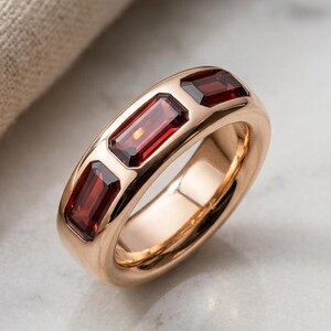 Può includere: Un anello in oro rosa con tre pietre preziose rettangolari rosso intenso. L'anello ha una finitura liscia e lucida e un design a fascia classico. Le pietre preziose sono incastonate in un'incastonatura a canale, creando un look elegante e moderno.