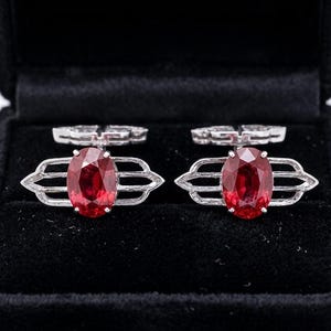 May include: A pair of silver-toned cufflinks featuring a central oval-cut deep red gemstone. The metal setting has a geometric, elongated frame extending horizontally from the stone, with a hinged toggle mechanism visible at the top of each piece.