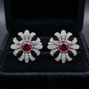 Puede incluir: Un par de gemelos de tono plateado con un diseño en forma de cruz, una piedra preciosa roja redonda en el centro y pequeños cristales transparentes. El metal tiene un acabado texturizado y las piezas están dispuestas simétricamente.