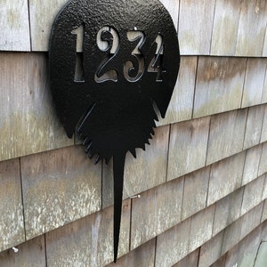 Pu&ograve; includere: Un cartello di numeri civici in metallo nero a forma di ferro di cavallo con il numero 1234 ritagliato. Il cartello &egrave; montato su una parete di legno.