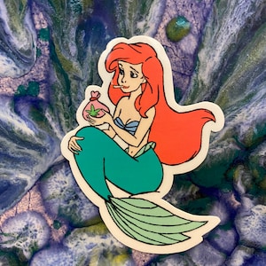 Puede incluir: Una pegatina con una sirena de dibujos animados con pelo rojo, un top azul y una cola verde. Sostiene una bolsa con un dise&ntilde;o de hoja de cannabis y un cigarrillo encendido. La pegatina est&aacute; sobre un fondo azul y verde.
