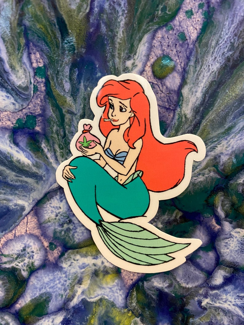 Puede incluir: Una pegatina con una sirena de dibujos animados con pelo rojo, un top azul y una cola verde. Sostiene una bolsa con un dise&ntilde;o de hoja de cannabis y un cigarrillo encendido. La pegatina est&aacute; sobre un fondo azul y verde.