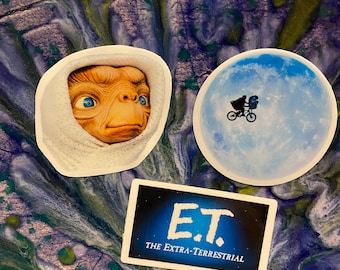 May include: Three stickers featuring the movie E.T. The Extra-Terrestrial. One sticker shows E.T. in his iconic white blanket, another shows E.T. riding a bicycle with a child, and the third sticker shows the movie title with a blue background.