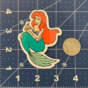 Puede incluir: Una pegatina troquelada con una sirena de dibujos animados con pelo rojo y cola verde, sosteniendo una bolsa con una hoja de cannabis. La sirena est&aacute; fumando un cigarrillo. La pegatina est&aacute; sobre un fondo de cuadr&iacute;cula azul.