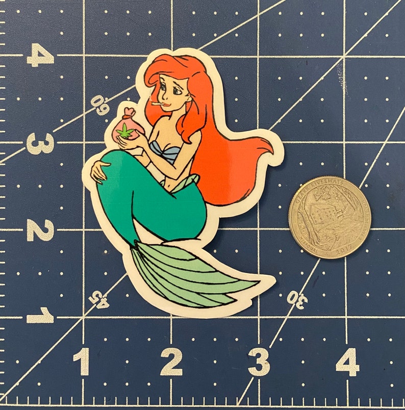 Puede incluir: Una pegatina troquelada con una sirena de dibujos animados con pelo rojo y cola verde, sosteniendo una bolsa con una hoja de cannabis. La sirena est&aacute; fumando un cigarrillo. La pegatina est&aacute; sobre un fondo de cuadr&iacute;cula azul.