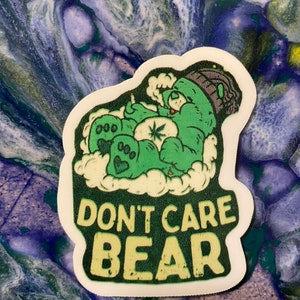 May include: A green sticker featuring a cartoon bear with a relaxed expression, surrounded by clouds. The bear is holding a bag with a cannabis leaf. The text "DON'T CARE BEAR" is displayed in a bold, yellow font.