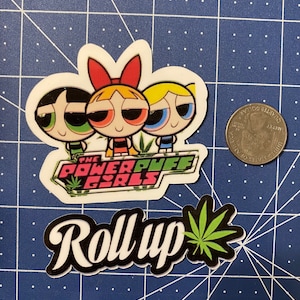 May include: A sticker featuring the Powerpuff Girls cartoon characters with the text "The Powerpuff Girls" and a green marijuana leaf. Below the sticker is a black and white sticker with the text "Roll Up" and a green marijuana leaf.