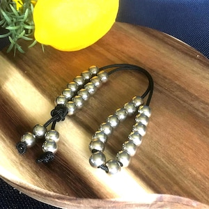 May include: Two silver beaded bracelets with black cords. The bracelets are on a wooden surface.