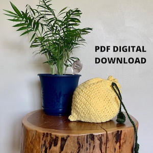May include: PDF digital download pattern for crocheting a yellow lemon-shaped drawstring bag with green drawstring. The bag is sitting on a wooden stump next to a potted plant.