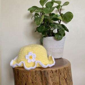 May include: A yellow crocheted sun hat with a white crocheted flower on top. The hat is sitting on a wooden stump with a green plant in a white pot behind it.