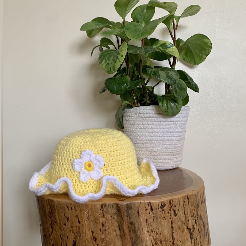 May include: A yellow crocheted sun hat with a white crocheted flower on top. The hat is sitting on a wooden stump with a green plant in a white pot behind it.