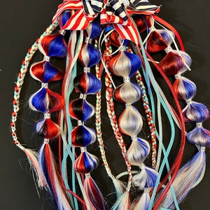 May include: A patriotic hair accessory in red, white, and blue. It features braided strands, decorative beads, and flowing ribbons. A large bow with red and white stripes and blue and white stripes adorns the top of the design.