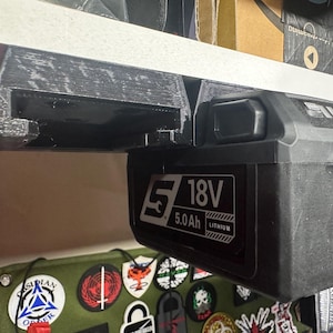 May include: Black 18V 5.0Ah Lithium battery pack with a black mounting bracket. The battery pack has white text that reads "18V" and "5.0Ah". The bracket is mounted under a white shelf.