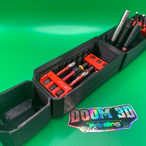 May include: Black 3D-printed tool organiser with red inserts, holding various screwdriver bits and tools. The organiser is open, revealing multiple compartments. The background is a solid green colour. The logo "DOOM 3D Printing" is visible.