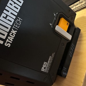 May include: Black rectangular storage container with the text "TOUGHBUILT STACKTECH" in white. Features an orange latch and silver accents. The container has an IP65 waterproof rating.